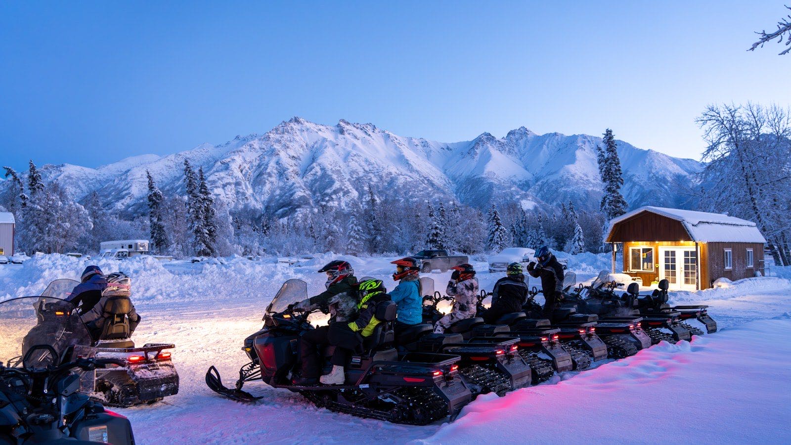 Group of touring snowmobiles lined up at dusk below big peaks